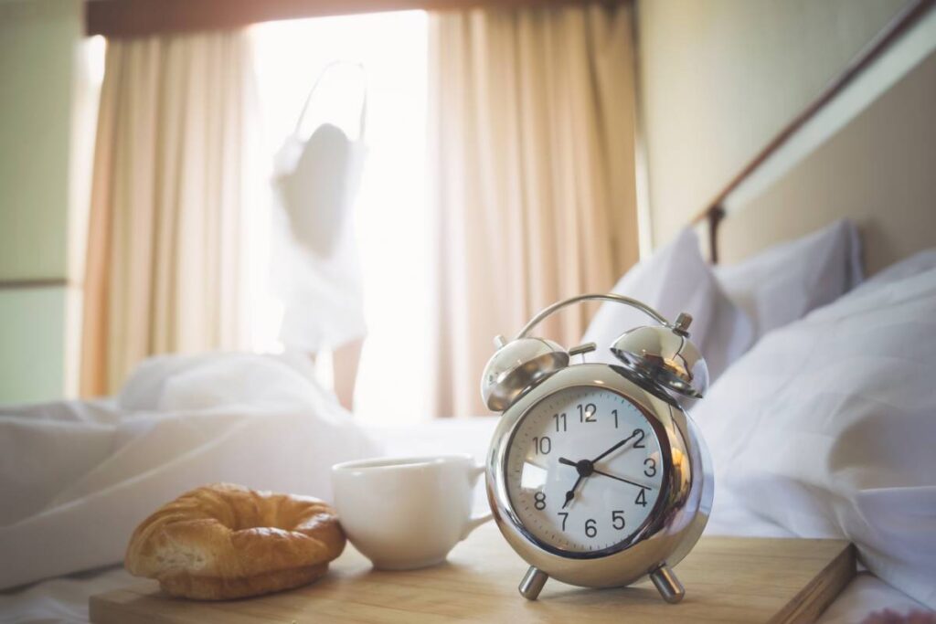 Alarm clock, cup of coffee and croissant having a good day with background happy woman stretching in bed after waking up, sunlight in morning.